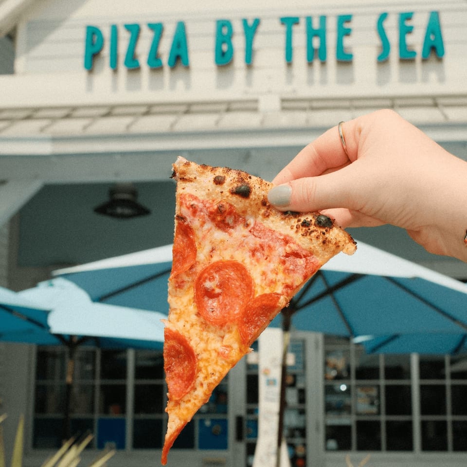 Hand holding a slice of pepperoni pizza in front of the "Pizza by the Sea" restaurant.