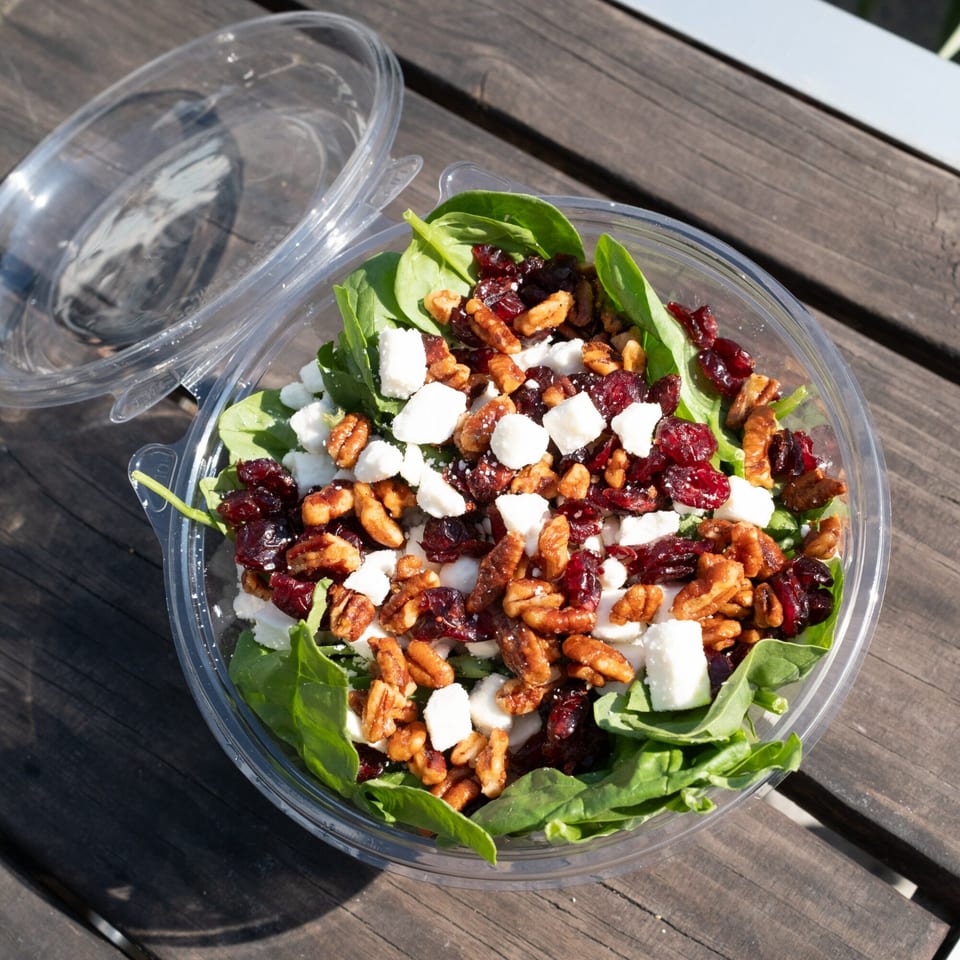 A spinach salad with cranberries, pecans, and feta cheese in a clear plastic container on a wooden table.