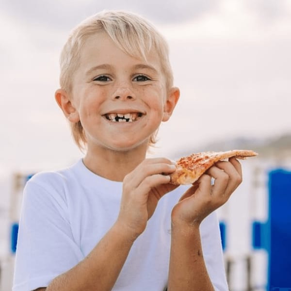 A smiling boy with missing front teeth holds a slice of Pizza by the Sea pizza.