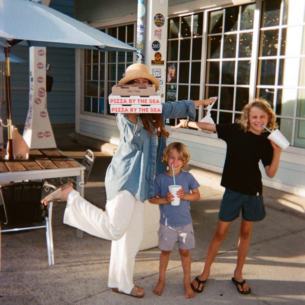 Woman in straw hat holding Pizza by the Sea boxes with two smiling boys drinking.