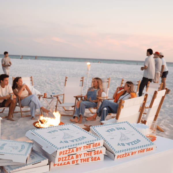 Pizza by the Sea boxes on a table with people around a bonfire on a beach at sunset.