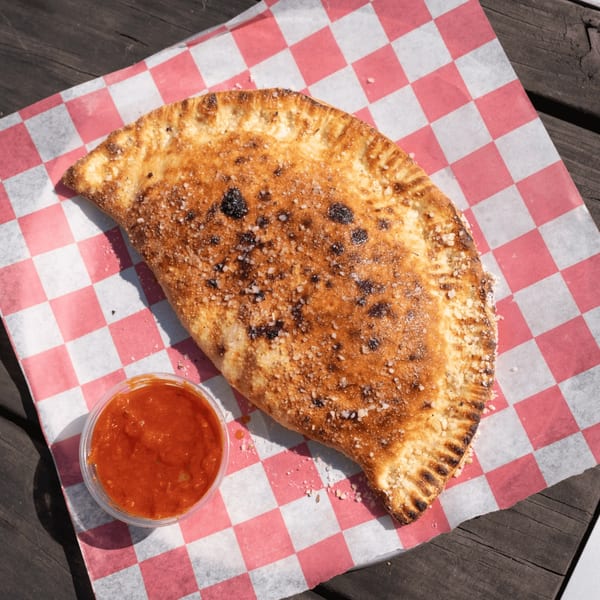 A golden-brown calzone with parmesan and a side of marinara sauce on red and white checkered paper.