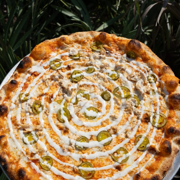 A Pizza by the Sea pizza with chicken, jalapeños, and a spiral of white sauce on a metal tray.