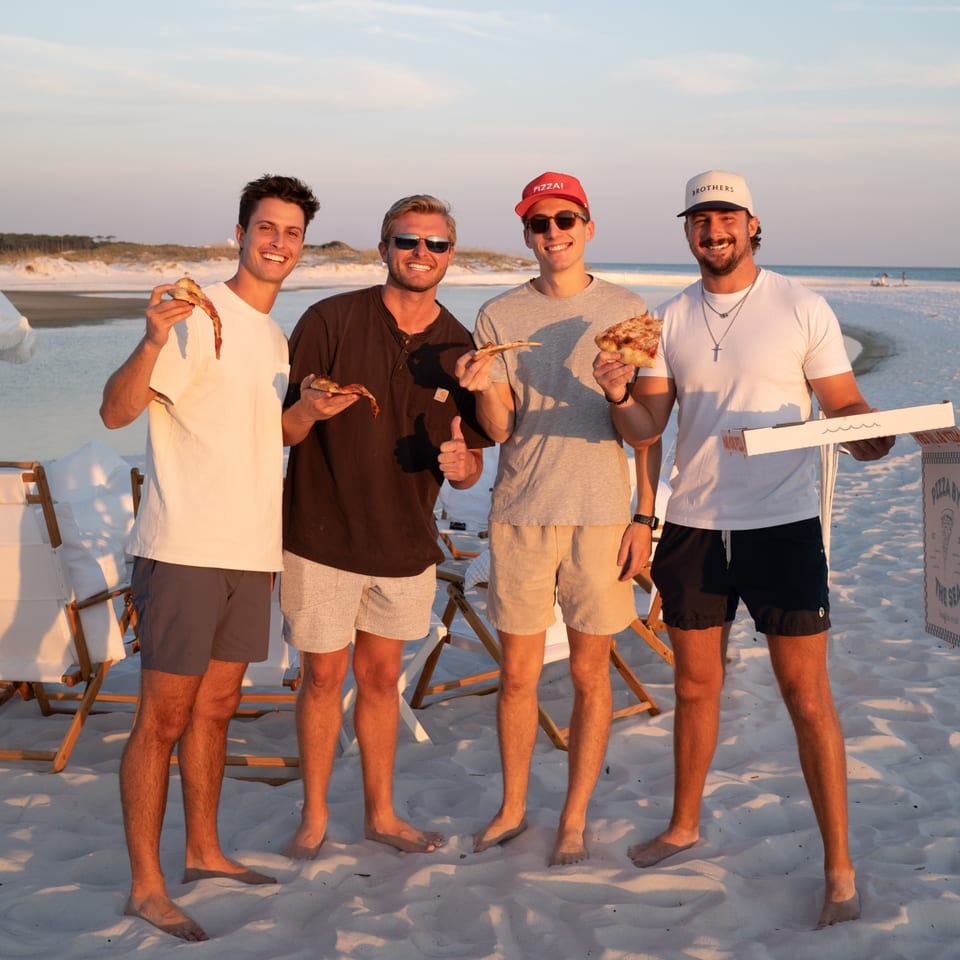 Four smiling men on a beach at sunset, holding slices of Pizza by the Sea pizza.