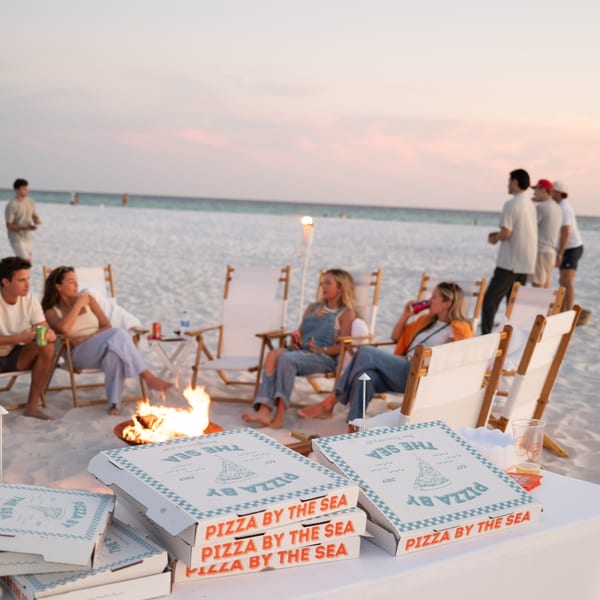 Pizza by the Sea boxes on a table with people relaxing around a bonfire on a beach at sunset.