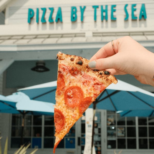 Hand holding a slice of pepperoni pizza in front of the "Pizza by the Sea" restaurant.