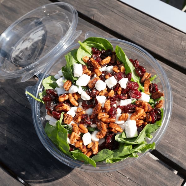 Spinach salad with cranberries, pecans, and feta cheese in a clear plastic container on a wooden table.