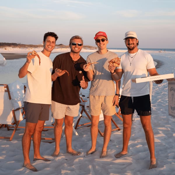 Four smiling men on a sandy beach, holding slices of Pizza by the Sea pizza at sunset.