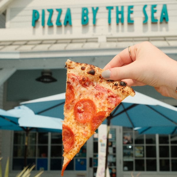 Hand holding a pepperoni pizza slice in front of the "Pizza By The Sea" restaurant sign.