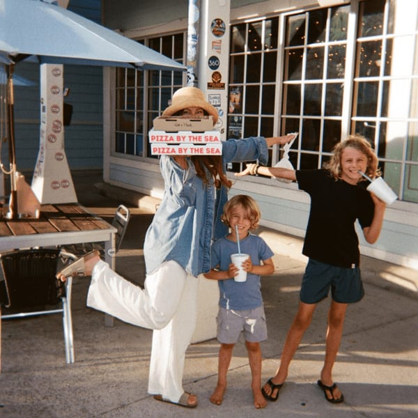 Woman holding Pizza by the Sea boxes with two children, all smiling outdoors.