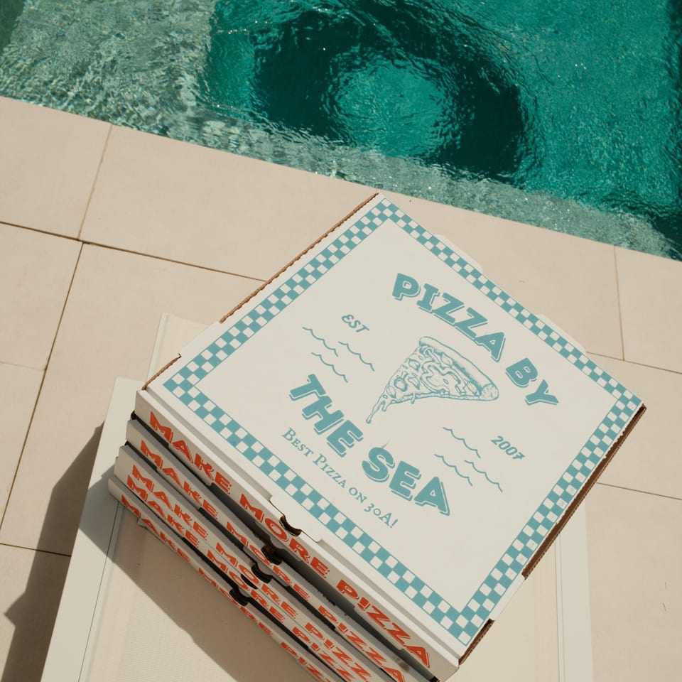 Stack of Pizza by the Sea boxes on a white lounge chair next to a swimming pool.